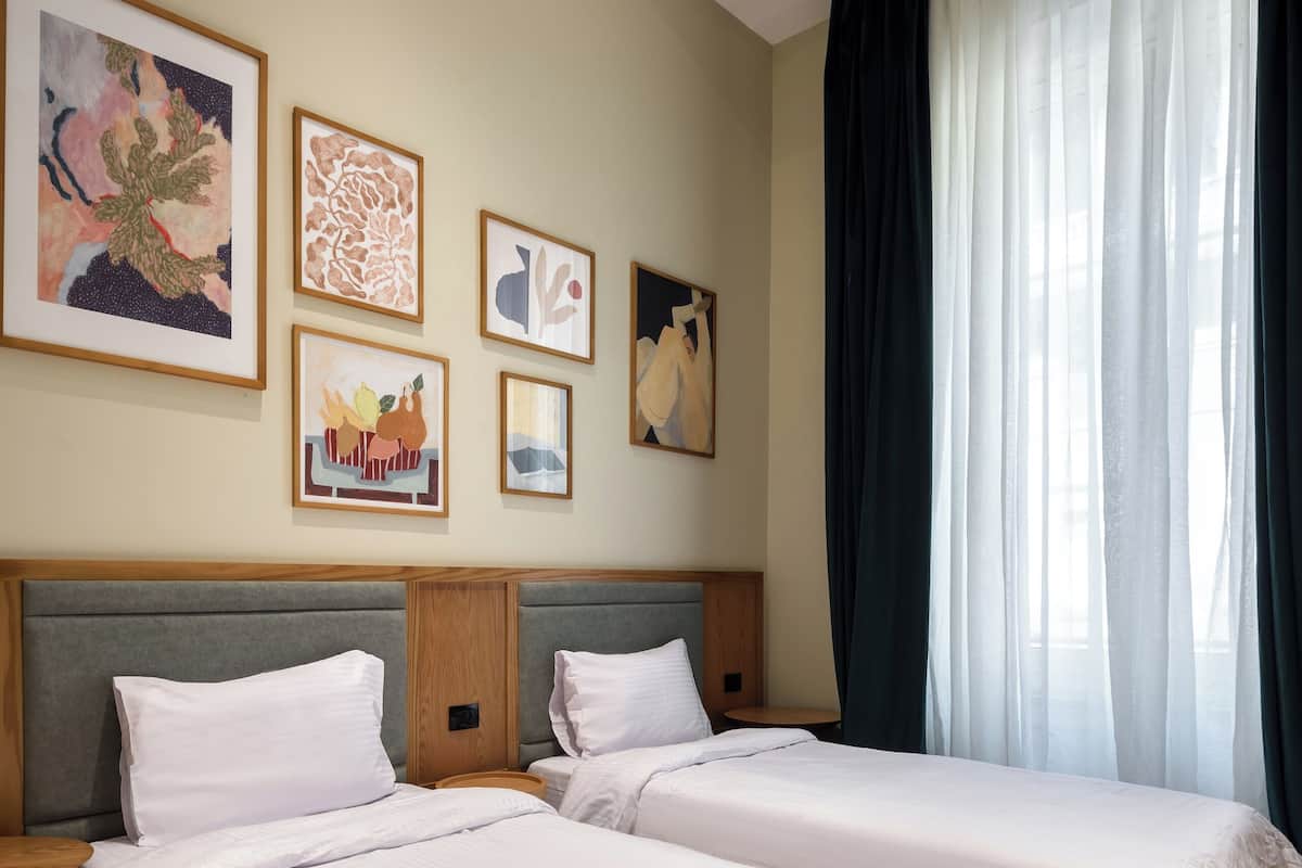 A hotel room with two beds, a large window with curtains, and artwork on the wall.