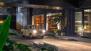 A modern building with a car parked in front, a covered entrance, and large green leaves in the foreground. - Wellesley Hotel (Pune)