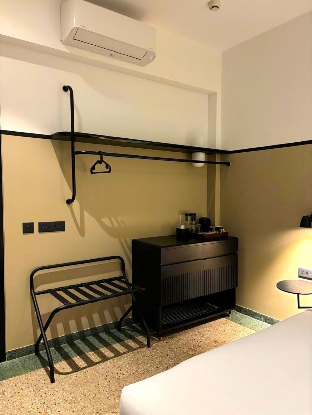 Habitación de hotel con cama, mesita de noche, una pequeña mesa, un estante metálico negro, un armario negro y un aire acondicionado instalado en la pared.