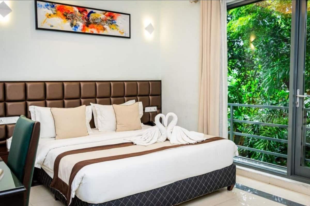 A hotel room with a large bed, a headboard, a desk, and a window with a view of greenery.
