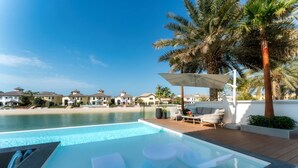 Piscina com espreguiçadeiras e uma palmeira, com vista para uma praia e algumas vilas.