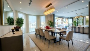 Uma área de jantar moderna com uma mesa comprida posta para seis pessoas, cercada por cadeiras. Há um espelho grande à esquerda, um lustre sobre a mesa e uma vista do exterior através de grandes janelas.