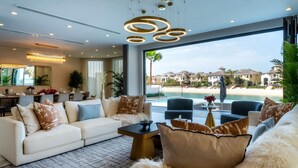 Uma sala de estar moderna com uma grande janela com vista para a praia e piscina, mobiliada com um sofá, poltronas e uma mesa de centro.