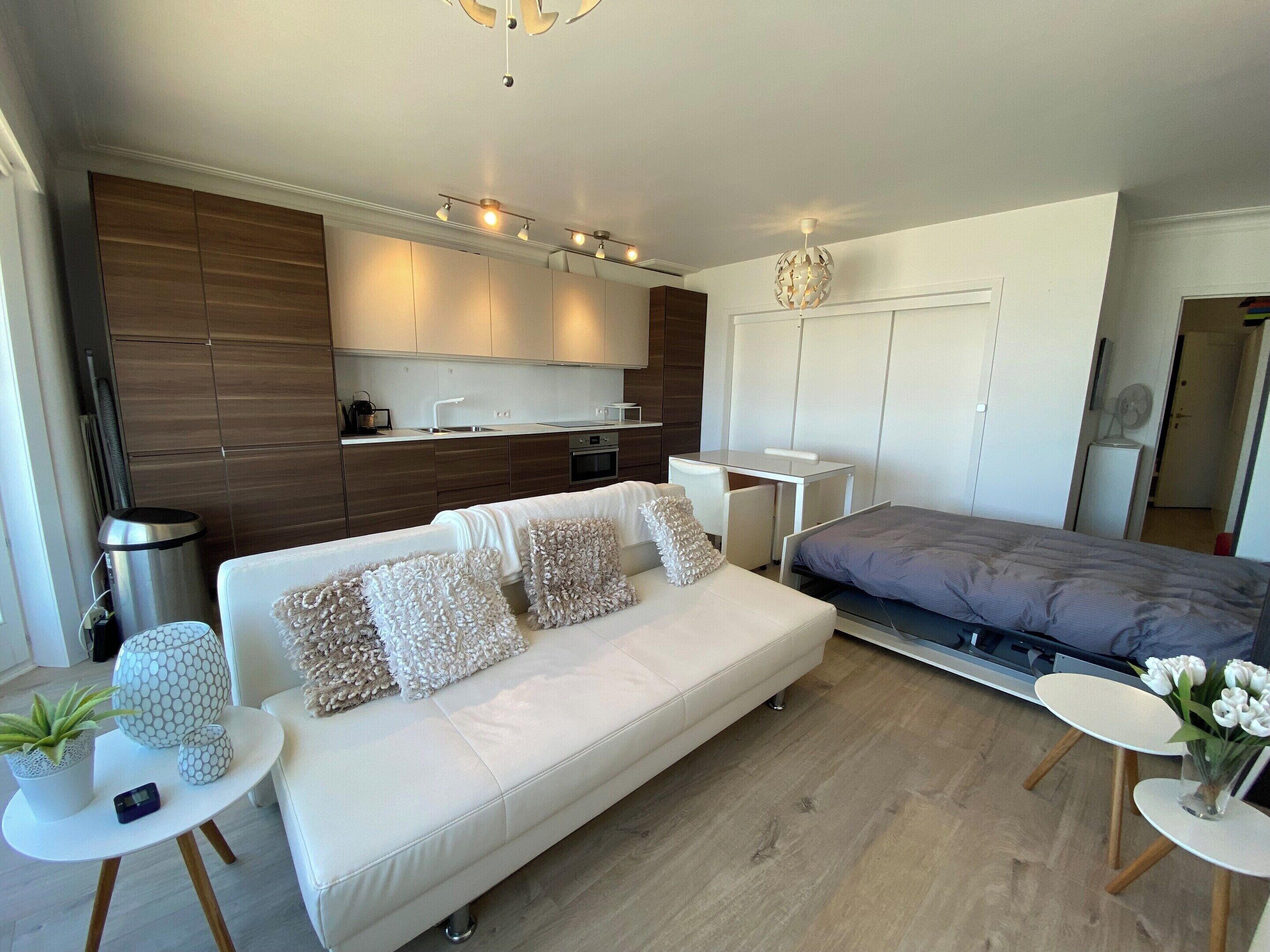A modern, compact living space with a white sofa, a small dining table, a kitchenette, and a bed.