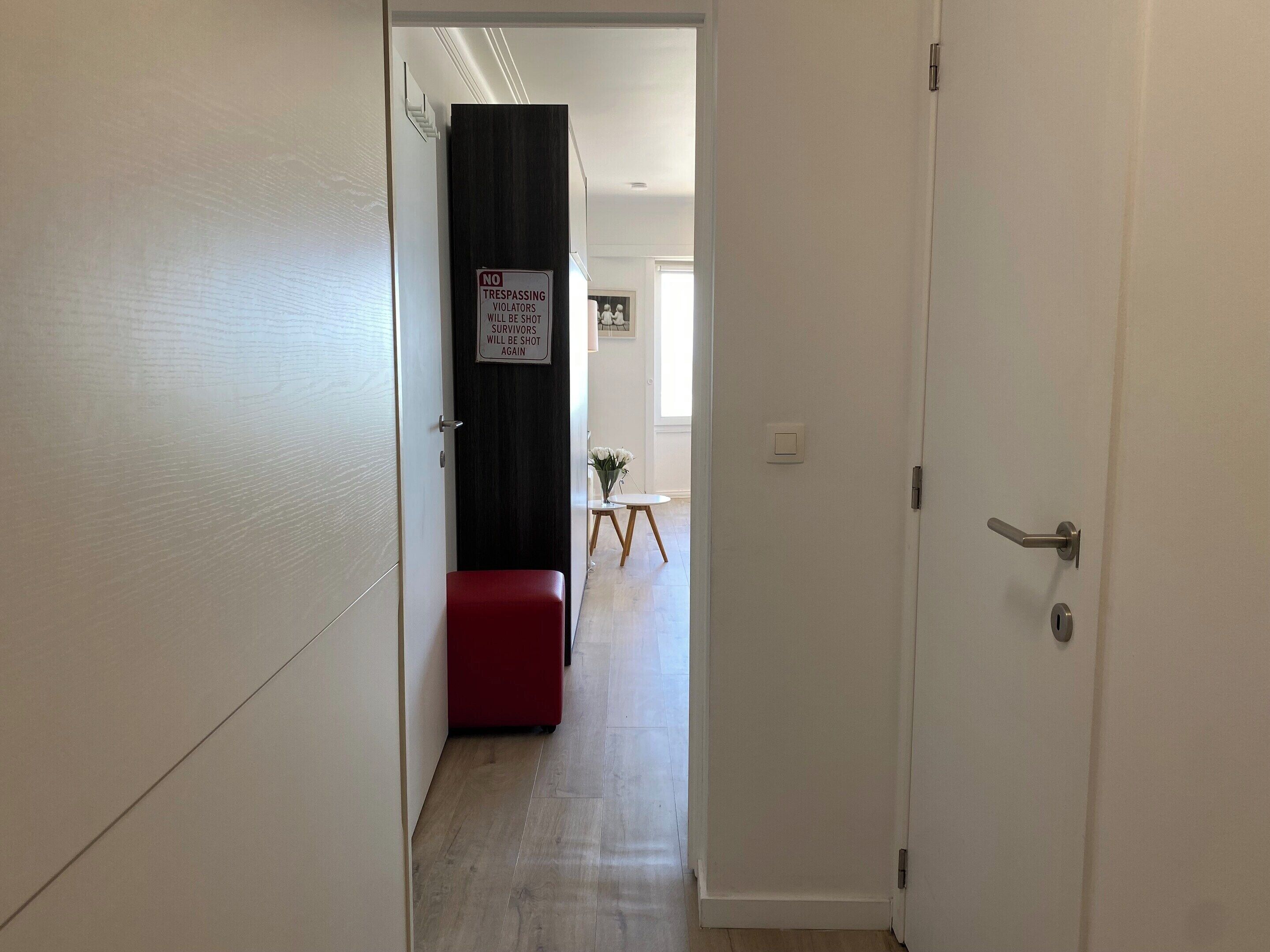 A hallway with a red bench, a door with a 'No Trespassing' sign, and a small table with flowers in the background.