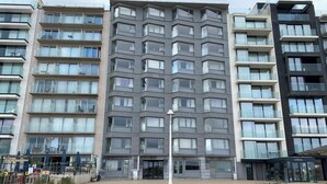 A modern multi-story building with balconies, a central entrance, and a paved plaza with planters. - Sea-view Apartment in Nieuwpoort (Nieuwpoort)
