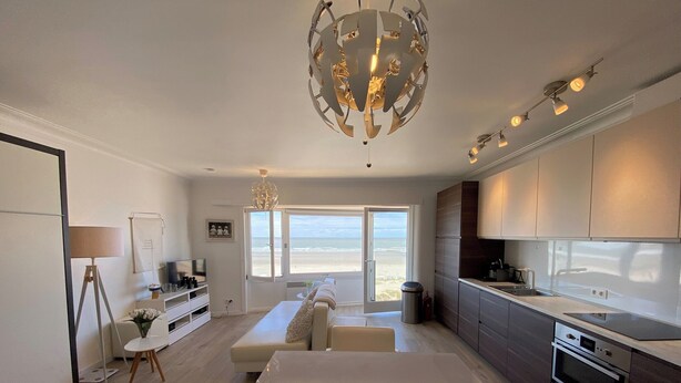 A modern kitchen with a large island, a dining area with a white table and chairs, and a view of the beach through a sliding door. - Sea-view Apartment in Nieuwpoort (Nieuwpoort)