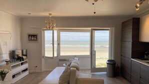 A modern living room with a sofa, a dining table, and a kitchen area. There are large windows with a view of the beach. - Sea-view Apartment in Nieuwpoort (Nieuwpoort)