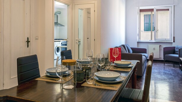 A dining area with a long table set for four, a sofa, and a window with shutters. - Corso Sardegna Design Apartment by Wonderful Italy (Genoa)