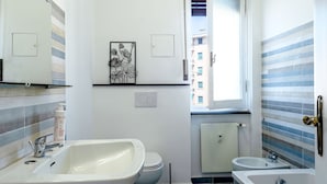 A modern bathroom with a large sink, toilet, and bathtub. There is a window providing natural light and a view of buildings outside. - Corso Sardegna Design Apartment by Wonderful Italy (Genoa)