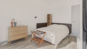 A bedroom with a bed, a wooden dresser, a wall-mounted shelf, and a floor lamp.