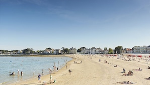 A beach with people sunbathing and swimming, a sandy shore, and buildings in the background.