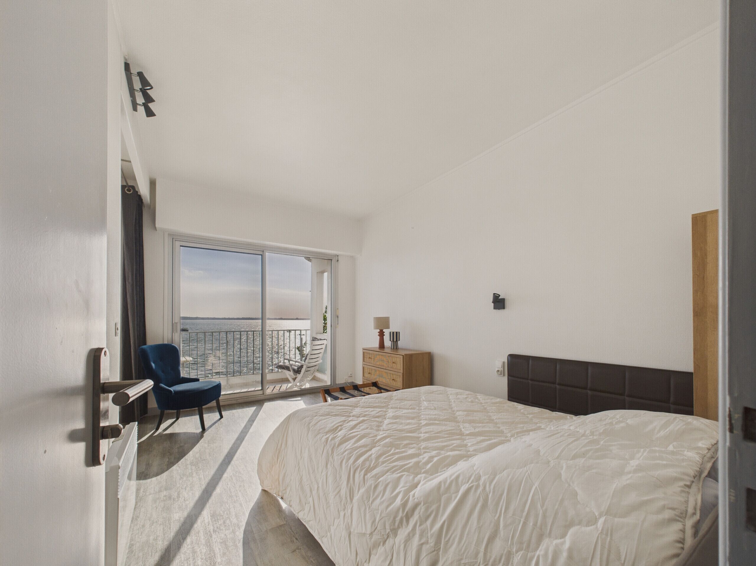 A bedroom with a large bed, a chair, a dresser, and a view of the sea through a sliding door.