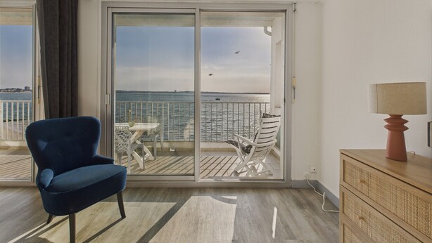 A balcony with a view of the sea, a blue chair, a white table, and a wooden dresser.
