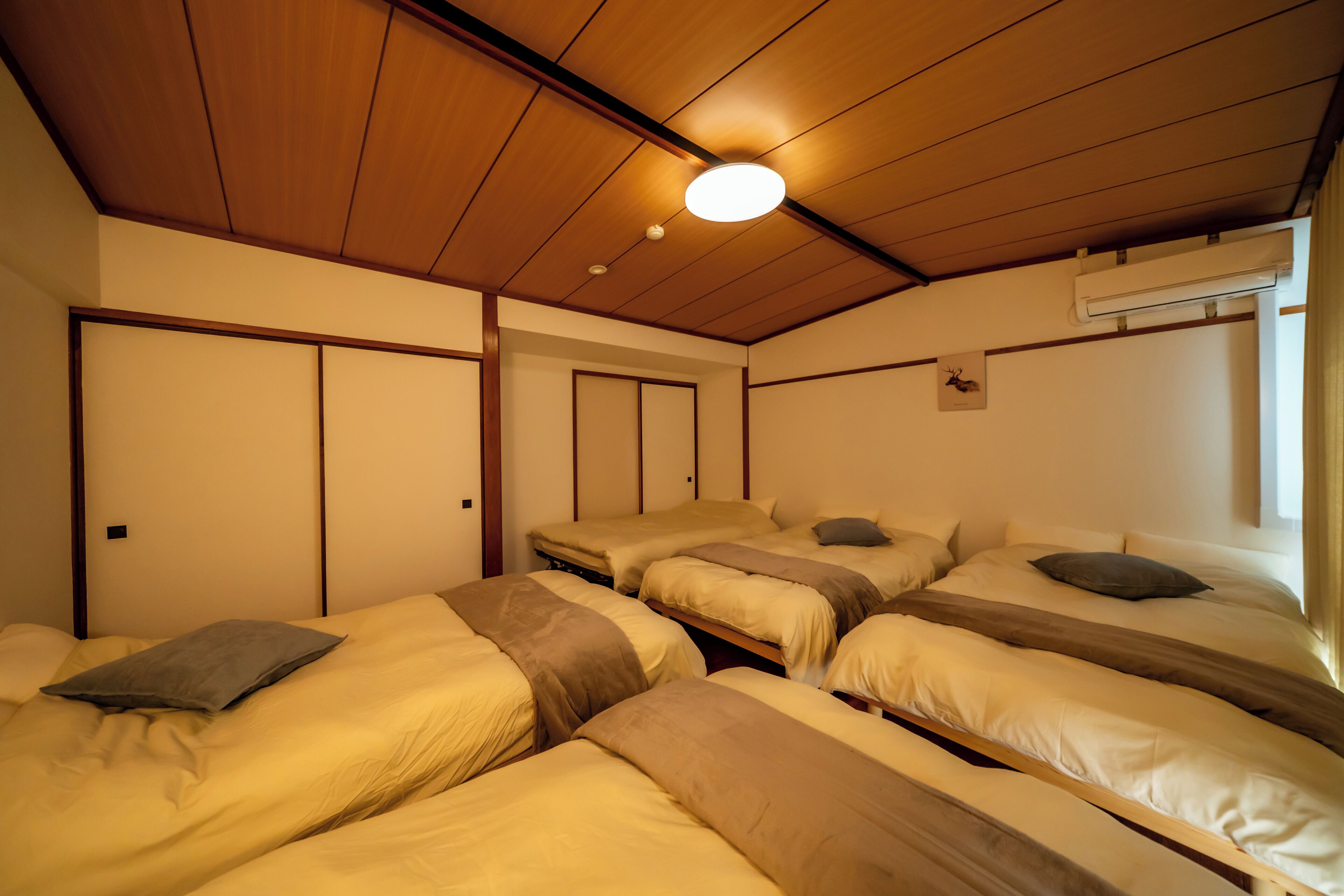 A room with three beds arranged in a row, wooden ceiling, and an air conditioning unit on the wall.