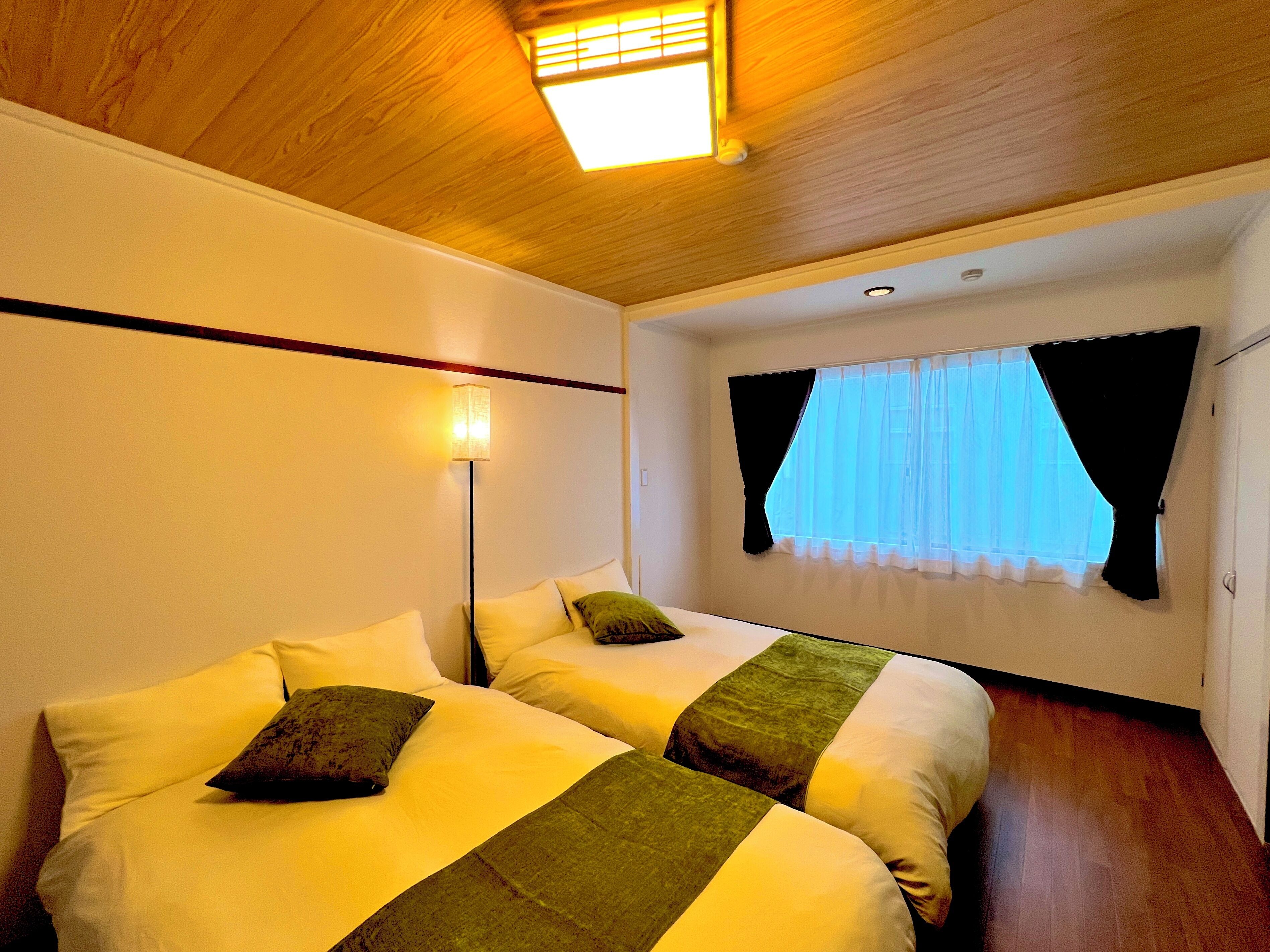 Ein Hotelzimmer mit zwei Betten, einer Deckenleuchte, einem Fenster mit Vorhängen und einem Holzboden.