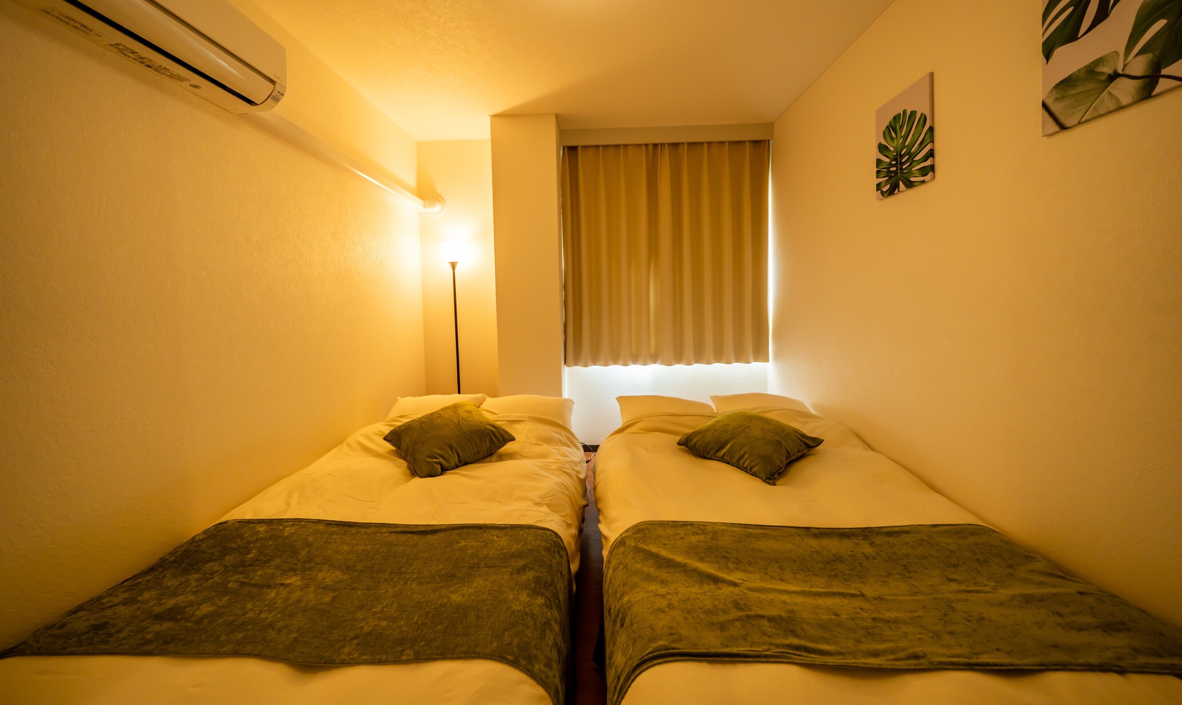 Een kleine, knusse kamer met twee eenpersoonsbedden, een bedlampje, een raam met gordijnen en een wandairconditioner.