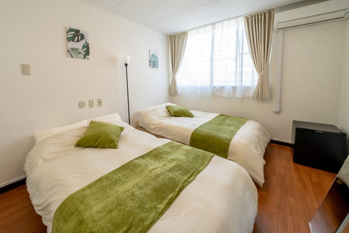 A hotel room with two beds, each with a green bedspread and pillow, white bedding, a standing lamp, and a black speaker.