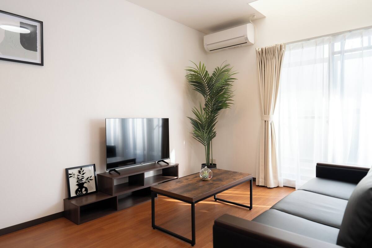 A modern living room with a flat-screen TV, a wooden coffee table, a sofa, a potted plant, and a large window with curtains.