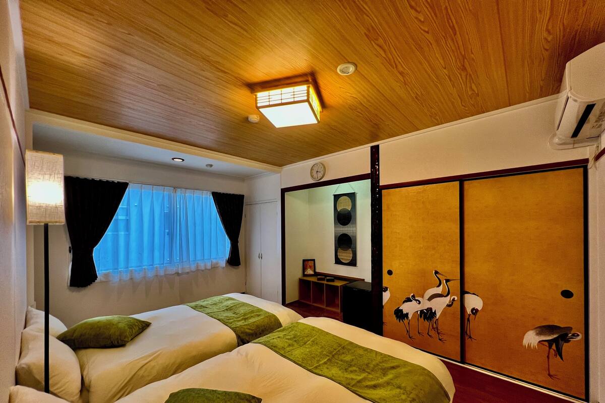 A hotel room with a wooden ceiling, a bed with green pillows, a window with sheer curtains, and a wall-mounted air conditioner.