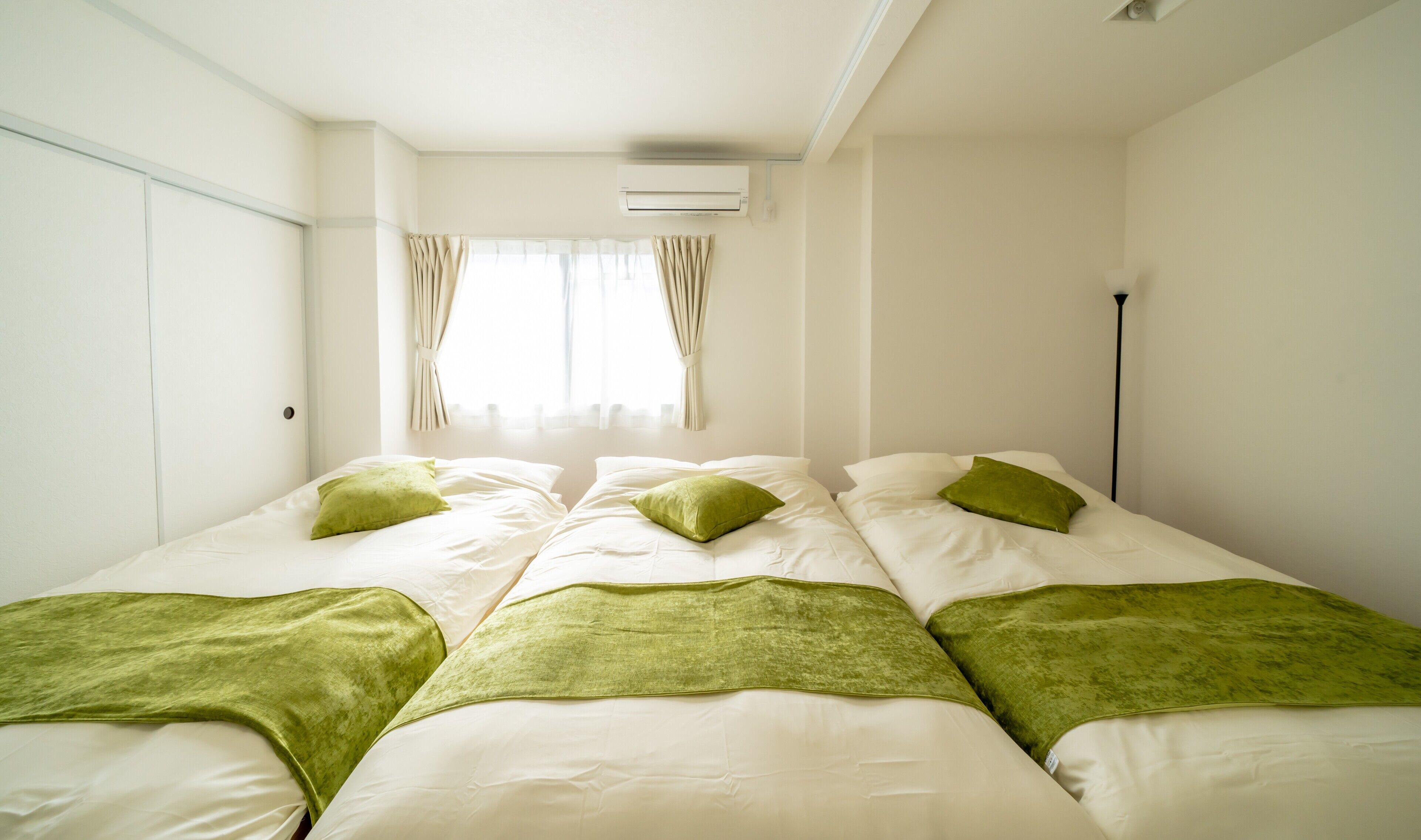 Habitación de hotel con dos camas, cada una con almohadas y manta verde. Se observa un equipo de aire acondicionado en la pared, una ventana con cortinas y una lámpara de pie.