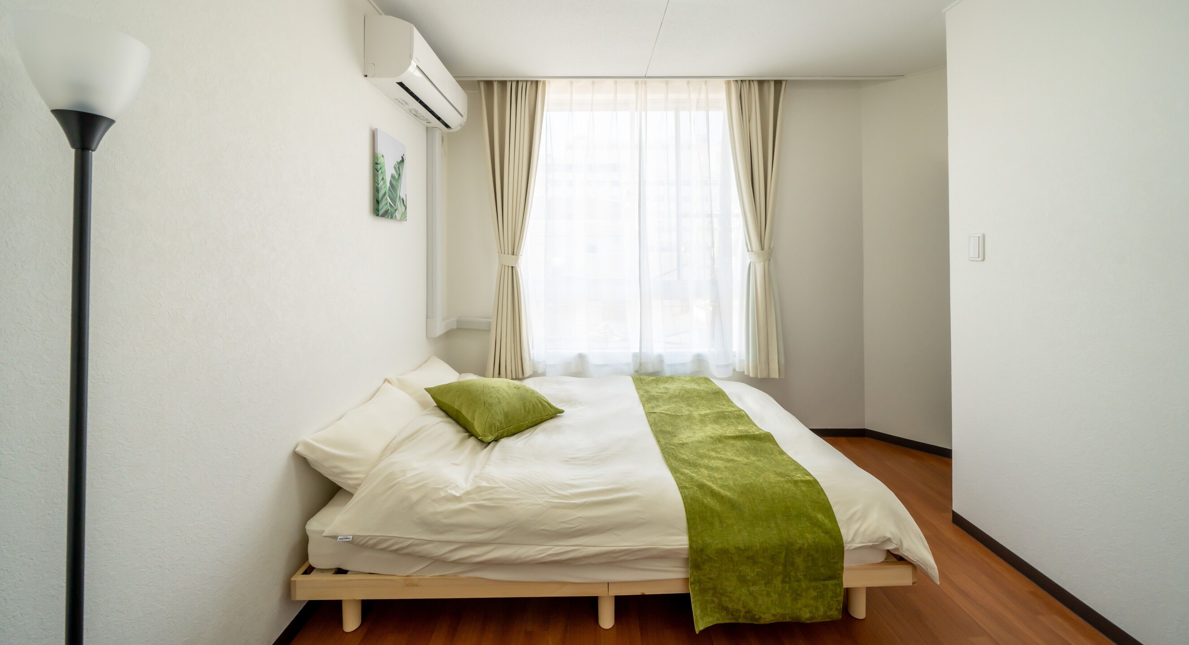 A bedroom with a bed, a bedside lamp, a window with curtains, and an air conditioning unit on the wall.