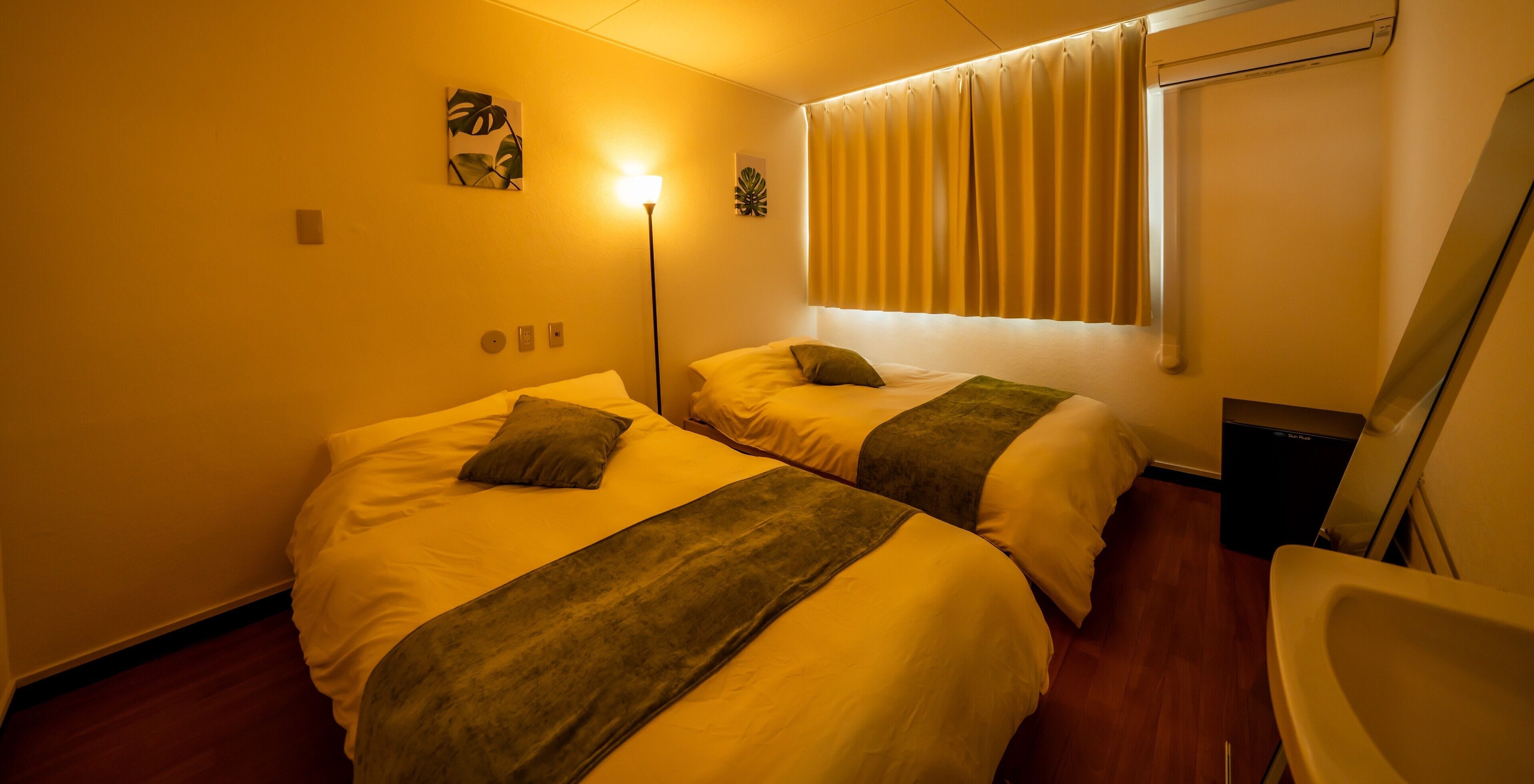 Une chambre d’hôtel avec deux lits, une lampe sur pied, une fenêtre avec des rideaux et une petite poubelle.