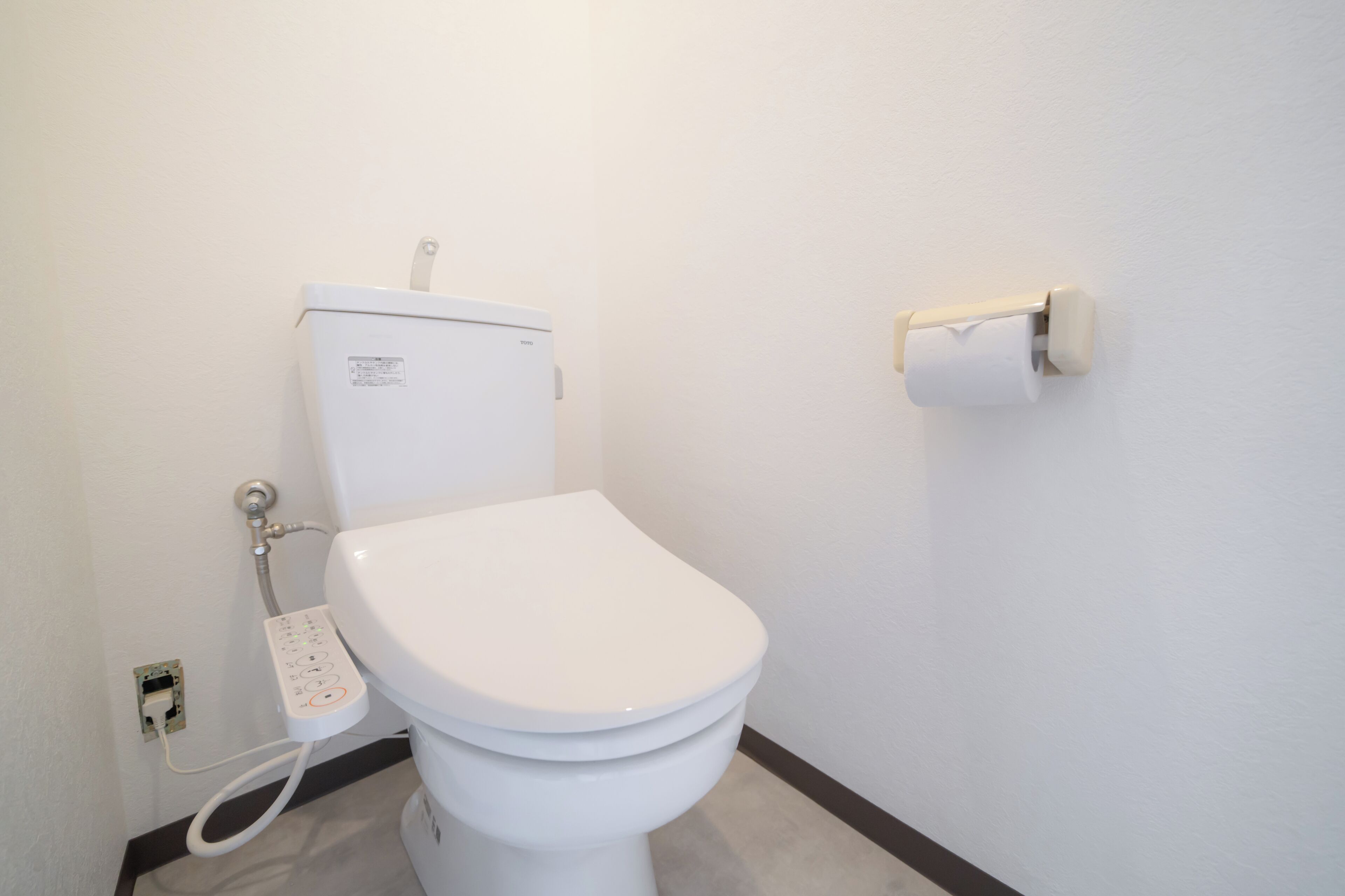 A white toilet with a control panel, a toilet paper holder on the wall, and a visible electrical outlet.