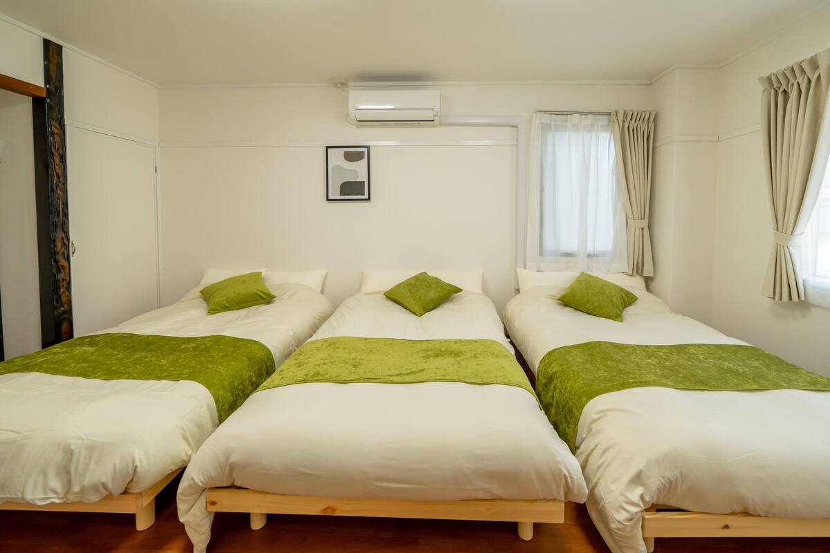 A room with three single beds, each with white bedding and green throw pillows. There is a ceiling air conditioner, a window with sheer curtains, and a framed picture on the wall.