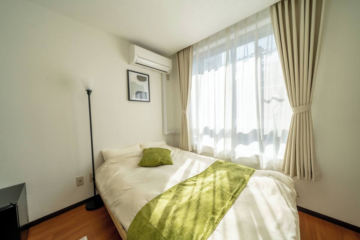 A neatly made bed with a green throw, a standing floor lamp, and a window with sheer curtains.