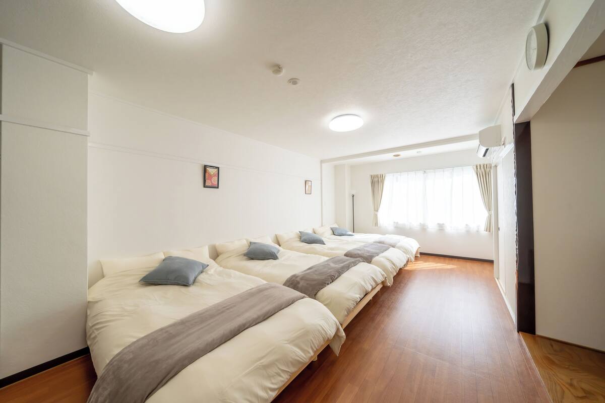 A hotel room with three beds aligned in a row, wooden flooring, white walls, and a window with curtains.