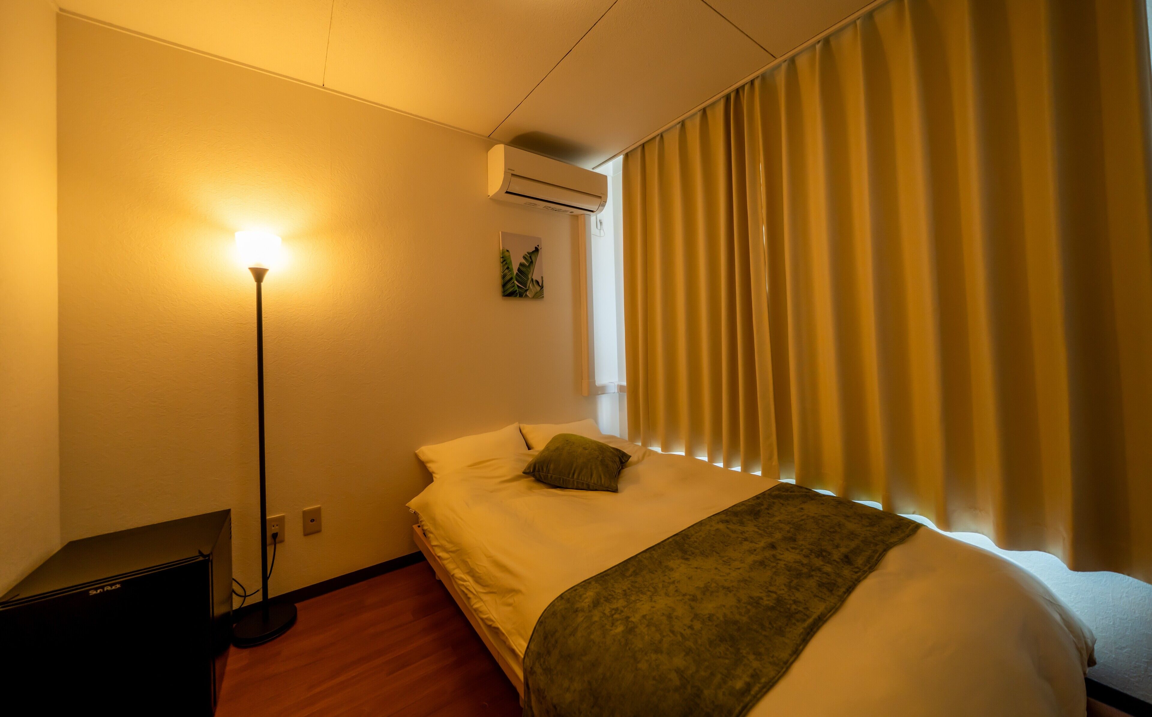 Una habitación de hotel con cama, lámpara de pie, aire acondicionado y ventana con cortinas.