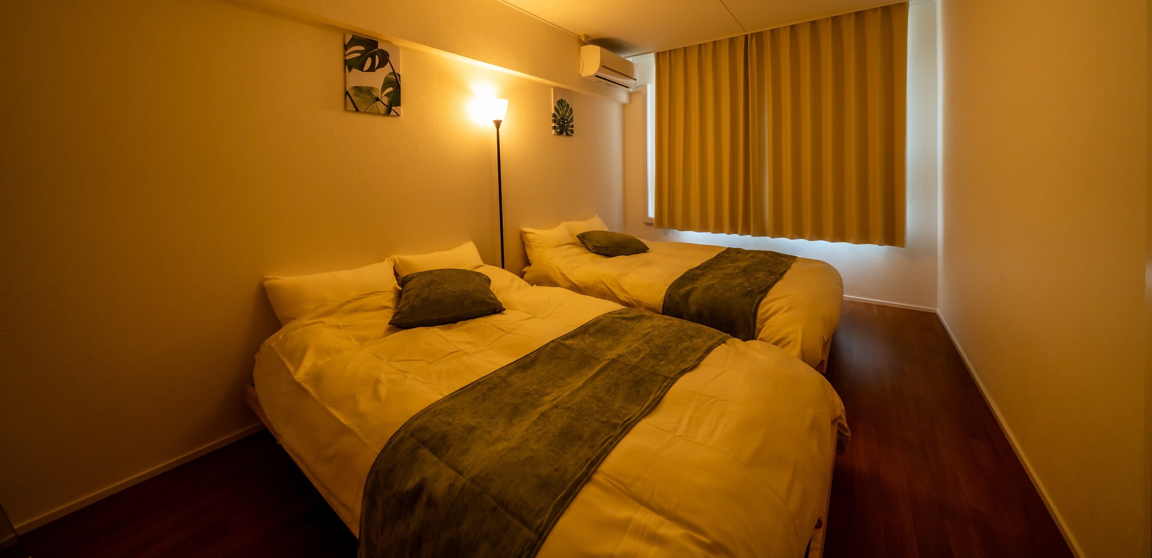 A hotel room with two beds, a standing lamp, an air conditioner, and a window with curtains.