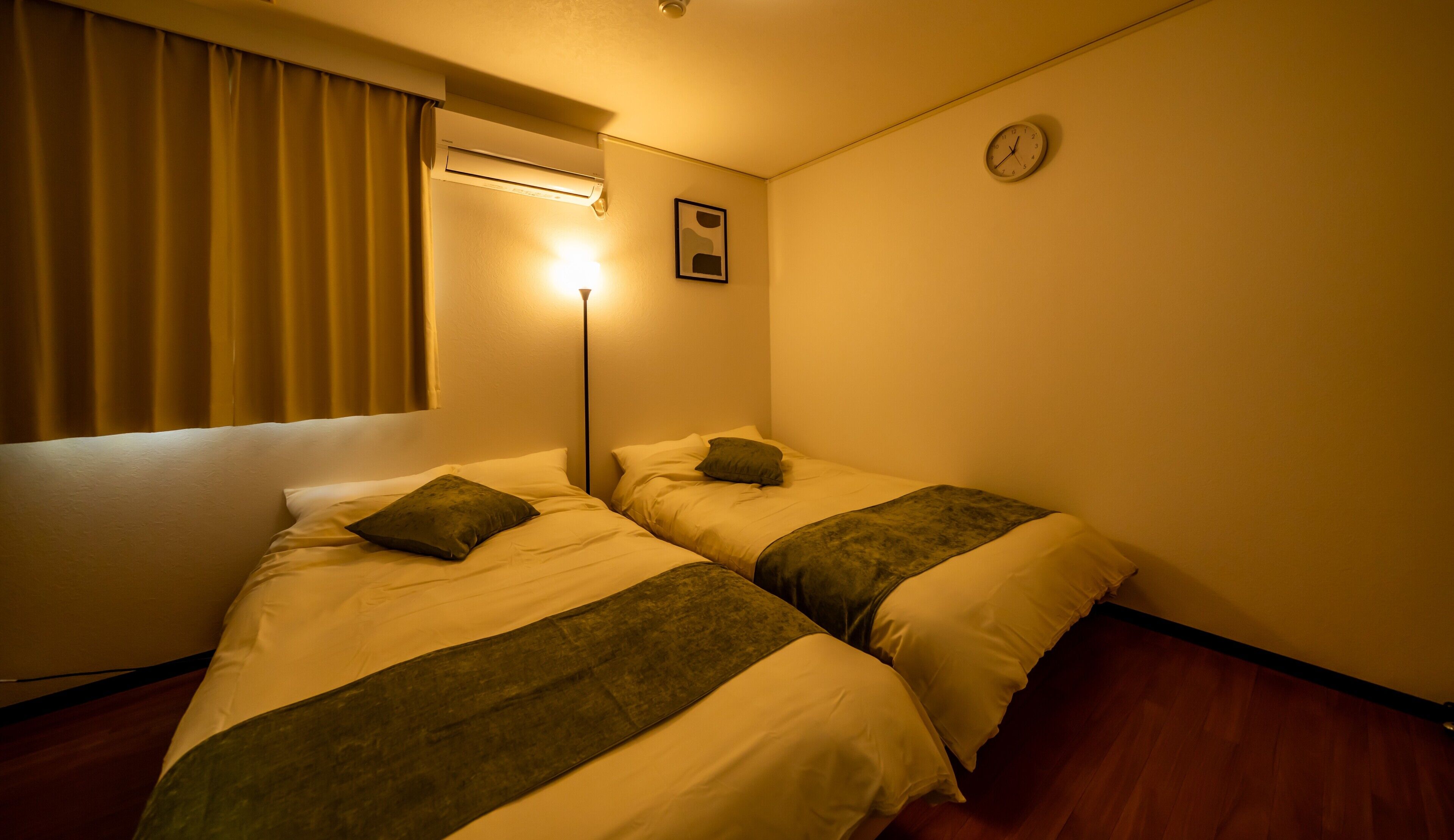 A hotel room with two beds, a standing lamp, a clock, and a window with curtains.