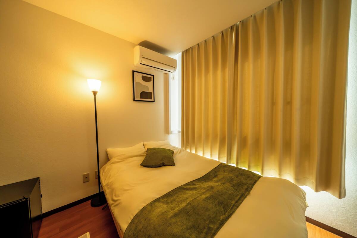 A hotel room with a bed, a standing lamp, an air conditioner, and a window with curtains.