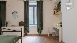 A room with a bed, a desk, a chair, a window with curtains, a mirror, and a shelf with plants.