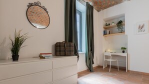 A room with a white dresser, a plaid suitcase, a potted plant, and a desk with a chair.