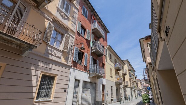 A narrow street with multi-story buildings, some with balconies and shutters, under a clear sky.