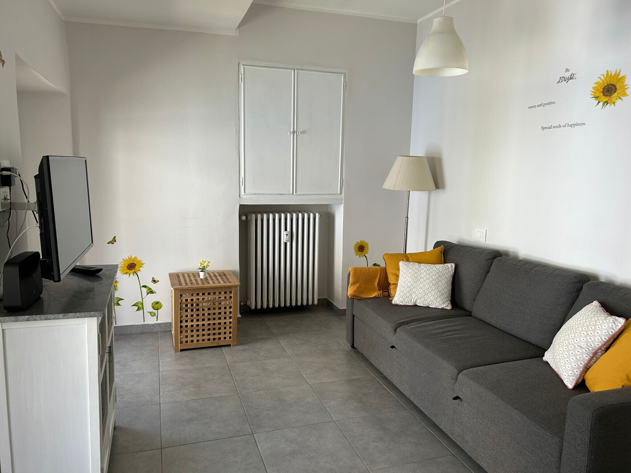 Ein modernes Wohnzimmer mit einem grauen Sofa, einem Fernseher, einer Heizung und dekorativen Sonnenblumen.