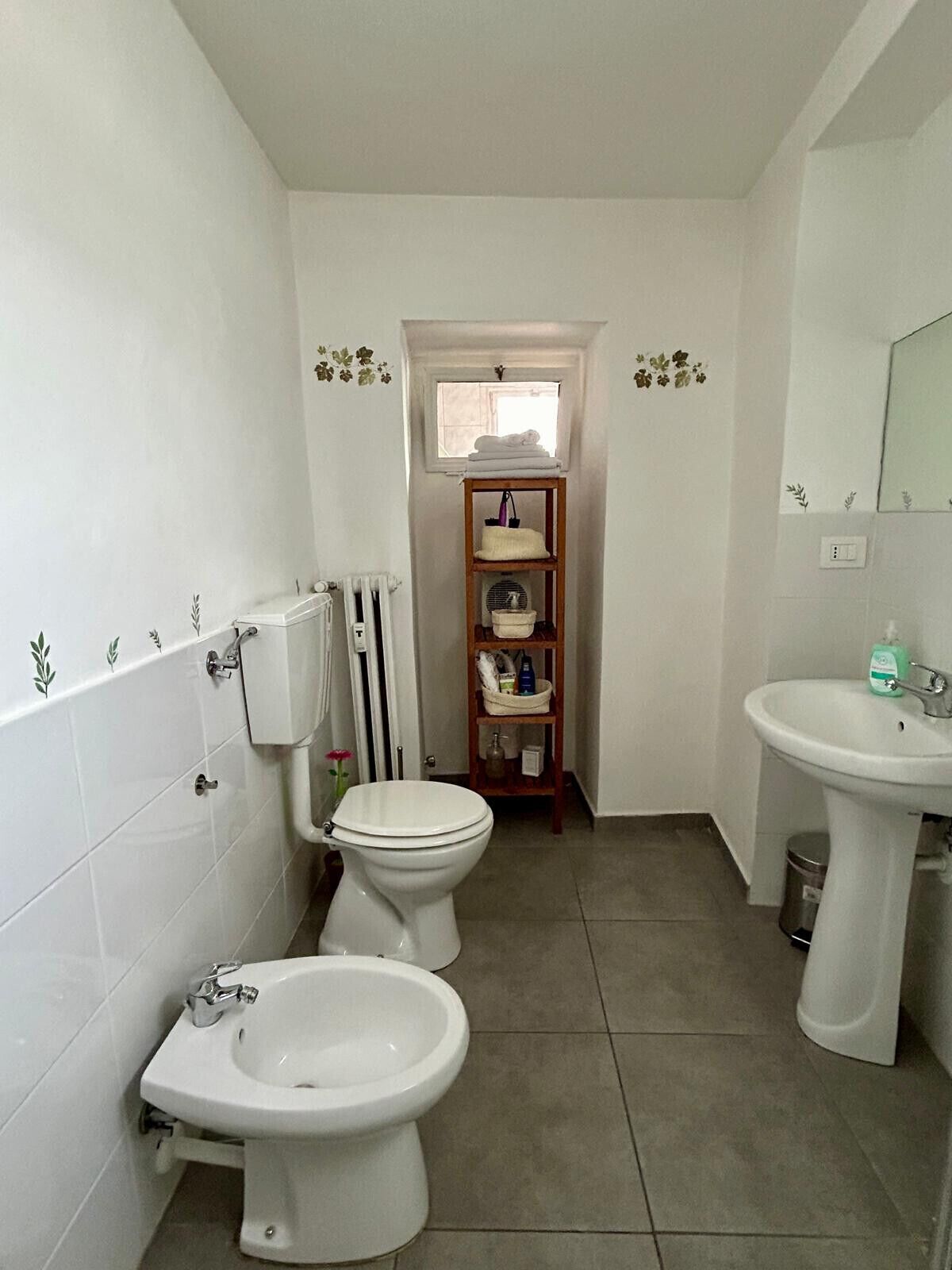 Een badkamer met een toilet, bidet en wastafel. Er is een plank met toiletartikelen en een raam boven het toilet.