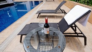 A round glass table with a bottle of wine and a glass, surrounded by two black lounge chairs with white cushions, near a swimming pool. - Astoria Apartment  (Nairobi)