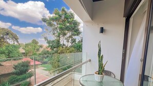 A balcony with a glass table and chairs, a potted plant, and a view of a residential area with trees and houses. - Astoria Apartment  (Nairobi)