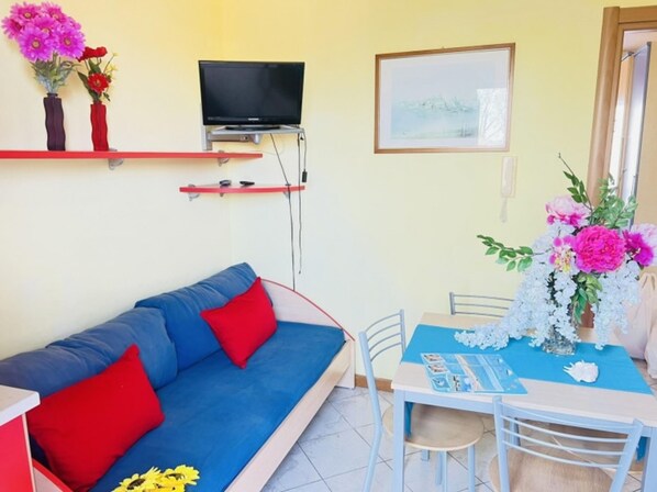 A room with a blue sofa, red cushions, a small table with a blue tablecloth, a TV on a stand, and a framed picture on the wall. - Colorful Flat in Bibione-beahost (San Michele al Tagliamento)