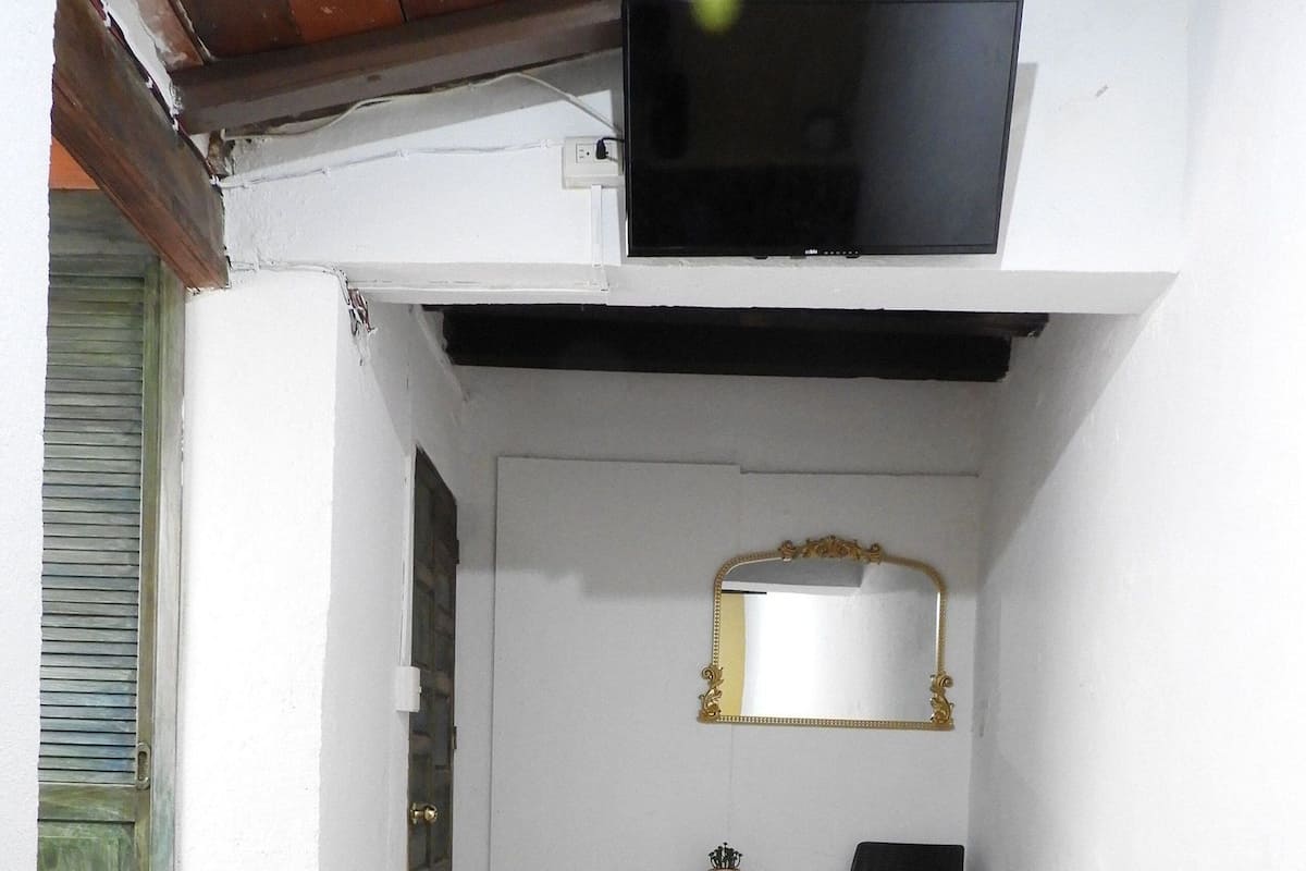A bedroom with a bed, a television mounted on the wall, a mirror, a chair, and a small table with a vase.