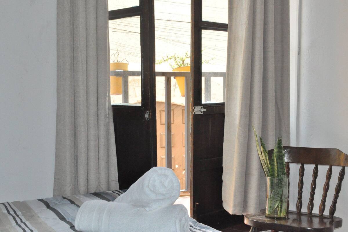 A room with a bed, a chair, a glass vase with plants, and a view through a door.