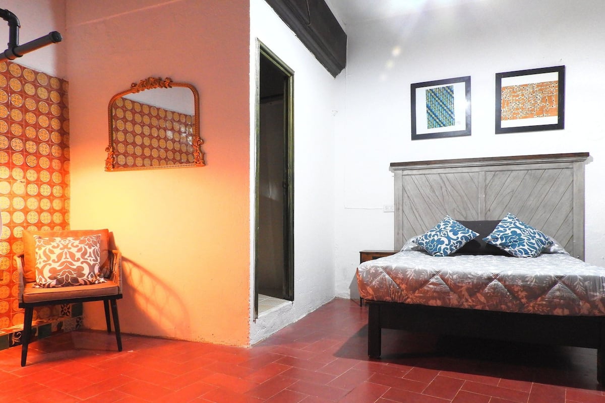 A bedroom with a bed, a chair, a mirror, and two framed artworks on the wall.