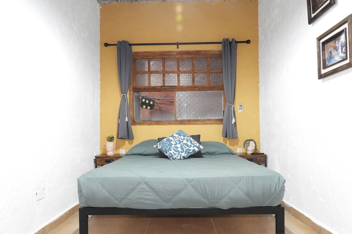 A bed with a quilted cover, a window with curtains, a small bedside table with a plant, and a wall-mounted picture frame.
