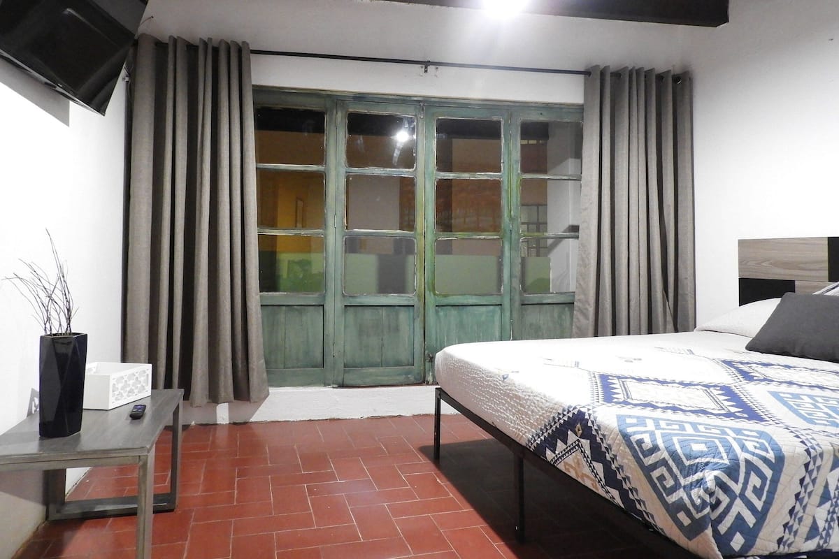 A bedroom with a bed, a TV mounted on the wall, a table, and a window with curtains.
