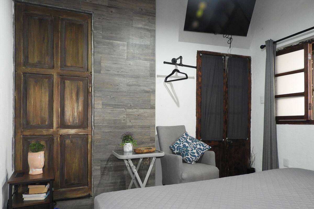 A bedroom with a bed, a chair, a small table with a plant, and a TV on the wall.
