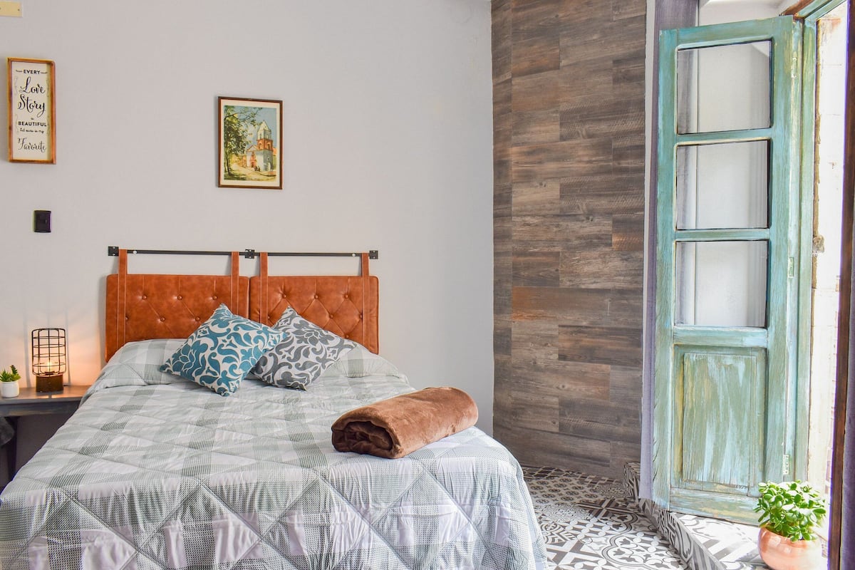 A bed with a patterned bedspread, a brown headboard, and a rolled towel on it.